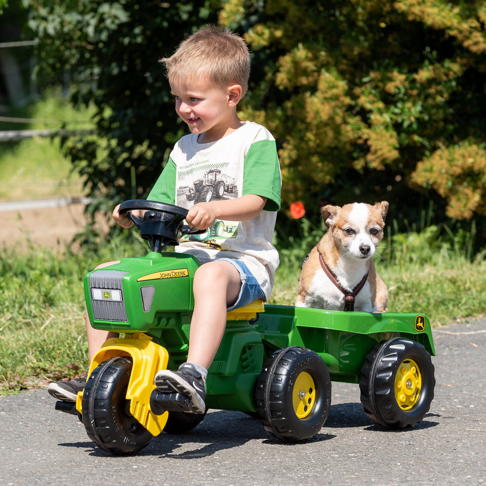 Lifestyle image of child riding on john deere three wheel tractor with dog in trailer 